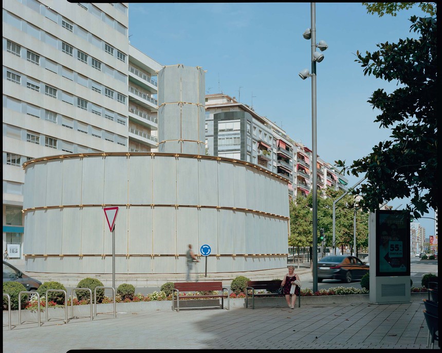 木板搭建的临时建筑立于圆形水池中央,周围为城市街道